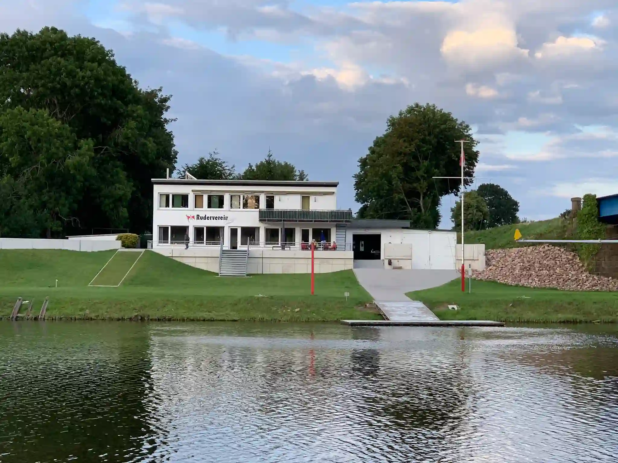 Erstellung des Hochwasserschutzes und Gestaltung der Aussenterrasse am Ruderhaus in Hoya 2020. Fotografie zeigt das Gebäude in Frontalansicht mit Nebengebäude, Hochwasserschutz und Terrasse samt Treppe von der Weser aus aufgenommen.